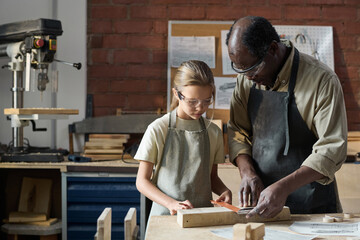 Waist up portrait of young girl measuring wood in workshop studio with senior carpenter helping