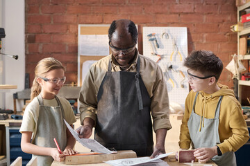 Waist up shot of two smiling children with senior carpenter looking at woodworking plans in workshop