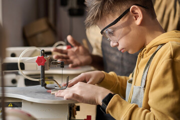 Side view portrait of focused Caucasian boy cutting wood using machine tools in carpentry workshop