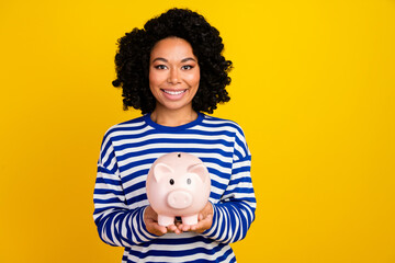 Fototapeta na wymiar Photo of good mood toothy beaming woman with chevelure dressed striped sweatshirt hold piggy box isolated on yellow color background