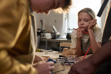 Portrait of young girl watching carpenter building wooden toys in crafting workshop for children