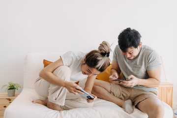 Asian Thai couple using mobile phone, sitting on white sofa in apartment room, both woman and man happy and enjoy playing game online, friend family spending time together.