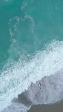 Vertical aerial top view of turquoise sea waves foaming and splashing, big waves from above rolling and breaking on empty ocean beach on cloudy day
