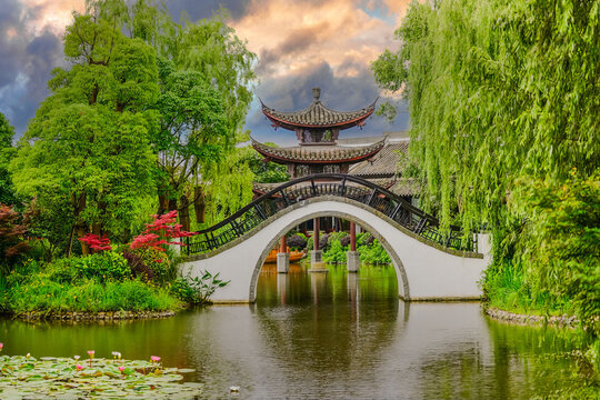 Traditional Chinese Garden with High-Arched White Bridge, Lush Greenery, and Serene Pond Reflections. - Powered by Adobe
