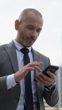 Vertical shot of happy bearded businessman in suit cheering celebrating success and achievement looking at his smart phone. Handsome professional man receiving good news in business