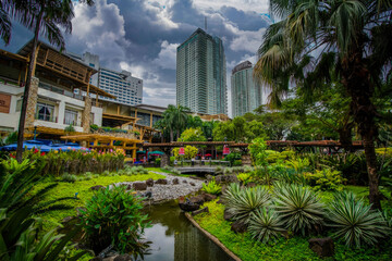 Fototapeta na wymiar Lush Urban Oasis with Tropical Plants and Stream Amidst High-Rise Buildings in a Modern City
