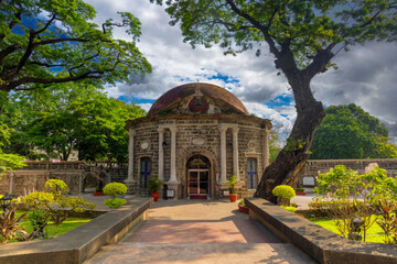 Naklejka premium Historical Circular Building with Dome Roof and Cross, Stone Facade, Arched Doorways, Lush Garden Pathway
