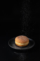 Single fresh baked donut sprinkled with falling sugar powder on dark retro plate on black...