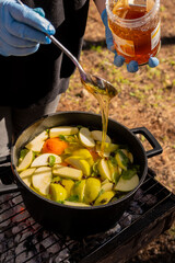 Golden honey being poured from a jar into a simmering pot of mulled wine with fruits on a grill, outdoors under sunlight