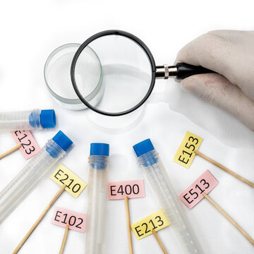 There is a magnifying glass in the laboratory assistant s hand, there is white powder on the laboratory table, next to there are signs with the names of laboratory food additives E and test tubes.