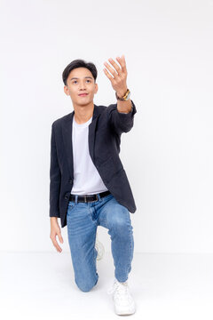 A charming young asian man kneels down, looking up and gesturing his hand up. Showing devotion. Front view, whole body shot, isolated on a white background.