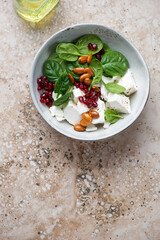 Bowl with feta cheese, baby spinach, pomegranate and almonds salad, above view on a beige granite background, vertical shot with space