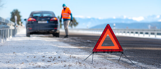 The warning triangle reflector on the road before a brokeded car in the background. Winter season on the road.