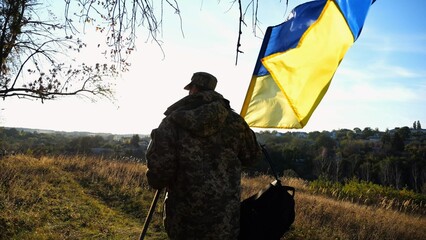 Young male military in uniform walks with waving flag of Ukraine at countryside. Soldier of...