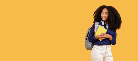 Smiling black student with notebooks looking to the side at free space on yellow background