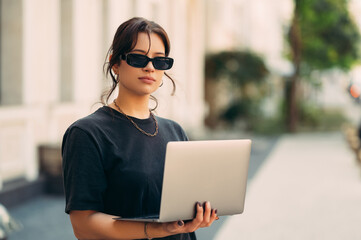 Young serious woman wearing sunglasses holds a laptop while standing outdoors on a summer day.