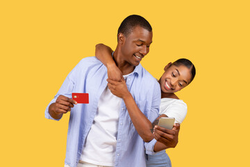 Delighted black couple shopping online, man with credit card, woman with smartphone, yellow backdrop