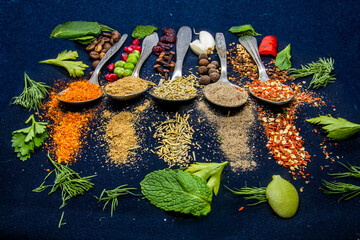 Five spoons with spices lie next to coffee, chili peppers and pomegranate seeds on a dark background next to peas, parsley, dill and green leaves very close	
