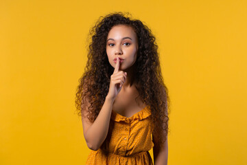 Smiling curly woman with finger on lips - shhh, secret, silence, yellow studio