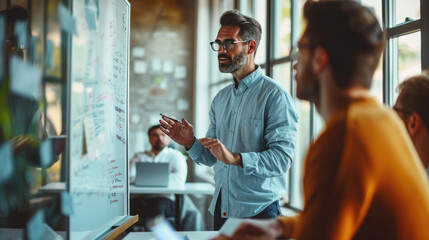 Man presenting or explaining something in front of a whiteboard with various colorful charts, diagrams, and notes, in a bright office setting