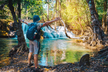 Back view of young man tourist enjoying in front of Huai mae khamin waterfall among rocks. Male traveler with backpack in tropical jungle in mountains overgrown with plants. Travel lifestyle concept.