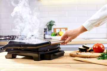 The woman's hands prepares meat on an electric grill on wooden table. Process of cooking meat on electric grill. Smoke in the home kitchen.