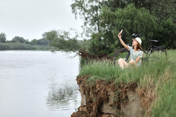 A beautiful cyclist makes a self-portrait using a smartphone. A young girl is resting on the river...