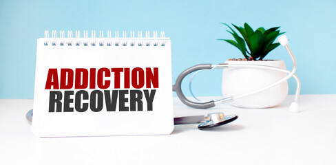 The text ADDICTION RECOVERY is written on notepad near a stethoscope on a blue background. Medical...