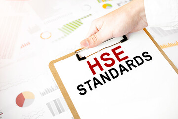 Text HSE STANDARDS on white paper plate in businessman hands with financial diagram. Business concept