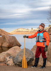 senior male paddler with a wooden canoe paddle on a rock shore of a mountain lake - Horsetooth...