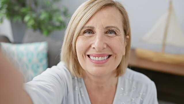 Caucasian mature woman with blonde hair smiles during a selfie at home showing joy and warmth.