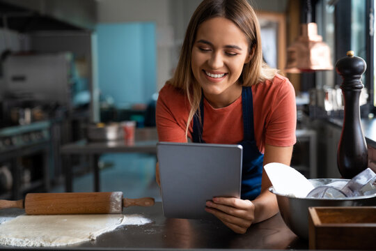 Smiling young woman baker using digital tablet at work in the pastry kitchen. Beautiful happy female baker looking at fintech device while working in the bakery.