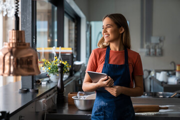 Young adult smiling female baker looking through window while standing in pastry kitchen and...