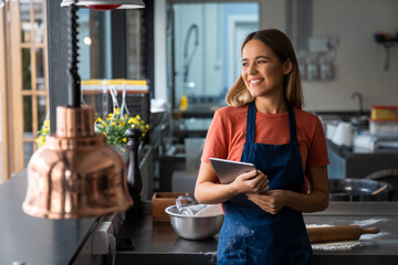 Successful woman baker wearing apron holding digital tablet pc looking through the window in bakery kitchen. Smiling blonde young woman with fintech device in pastry kitchen.