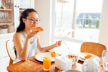 Young attractive Asian woman in glasses and casual clothes is sitting at table and eating sandwich....