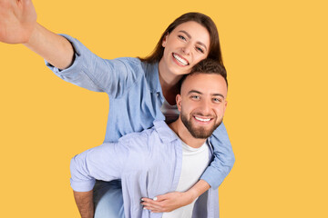 Happy loving european couple taking selfie together, cheerful young woman piggybacking her boyfriend, yellow background