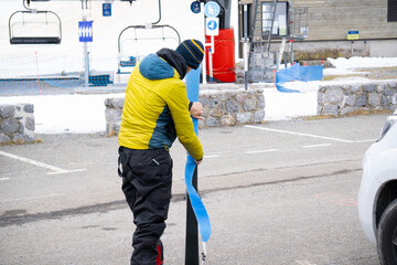 man putting sealskin on skis in parking lot, next to camper van, ski lift in the background.