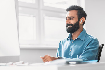 Smiling indian man with earbuds working at bright office desk