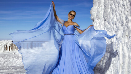 Fototapeta na wymiar Beautiful elegant woman wearing blue fluttering long dress and is posing on a sunny day in the mountains. Snow queen.