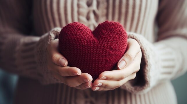 Close-up of a woman in a warm sweater holding a knitted burgundy heart in her hands. Valentine's Day greeting card. A symbol of love.
