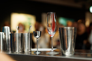 A wooden counter with a champagne flute and silver bar tools in the foreground and a blurred view...