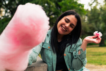 beautiful latin woman sitting, smiling and eating her cotton candy, enjoying the day, in an outdoor park