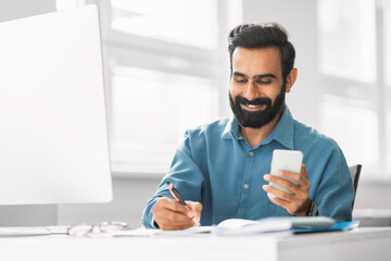 Smiling indian businessman using smartphone while writing notes