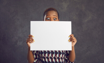 Funny surprised black woman showing empty white paper banner standing against grey background....
