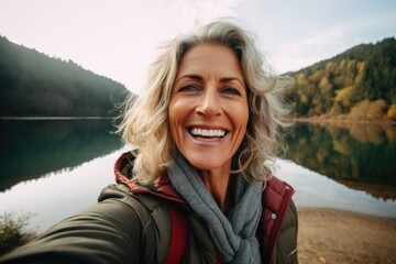 Smiling middle aged woman taking selfie lakeside - obrazy, fototapety, plakaty