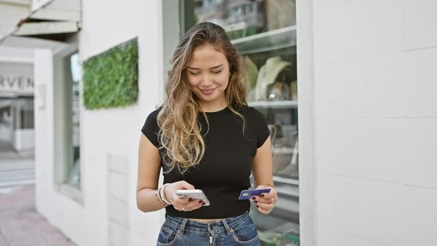 Confident and happy young hispanic woman enjoys outdoor shopping on the city street, using credit card and smartphone for carefree online payment with a beautiful smiling expression of satisfaction