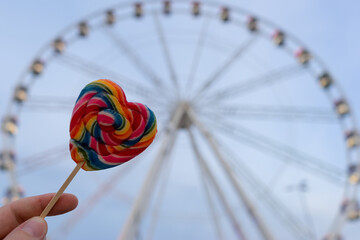 a multi-colored heart-shaped lollipop on the background of a Ferris wheel.