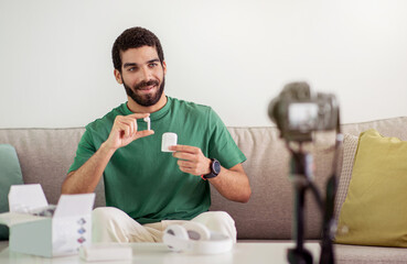 A cheerful young man in a green t-shirt sits on a couch, unboxing and showing a small electronic...