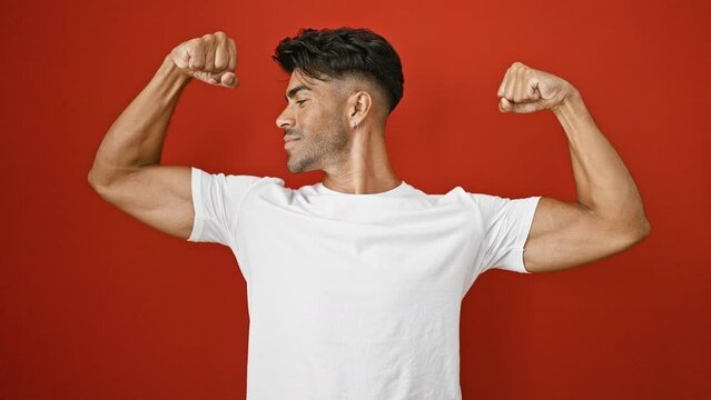 Hispanic man in white shirt posing with muscle flex against red wall exuding confidence.