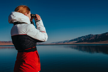 Young woman tourist standing on shore of lake and looking through binoculars in winter